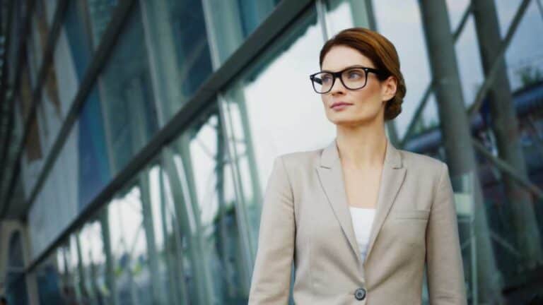 Portrait shot of the caucasian pretty business lady smiling to the camera and crossing her hands while standing on the glass building background outside